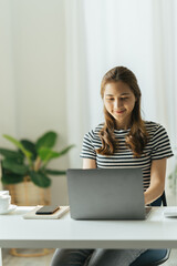 Fototapeta premium Smiling young beautiful Asian woman sitting working laptop computer at home.