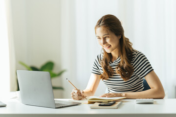 Young women take notes in their home office while working on a laptop.