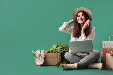 Happy young woman with laptop, credit card and new purchases on color background. Online shopping
