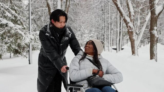 Young couple of asian man and his black girlfriend in the wheelchair talking on the snowy path