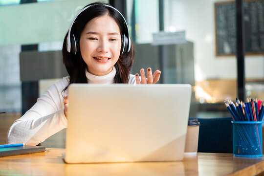 Female manager of Asian ethnicity who is happy in headphones looking at laptop screen. Chat happily with customers online. work remotely in the workplace education concept work-study at a coffee shop