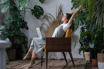 Fototapeta na wymiar Work-life balance. Happy female freelancer with closed eyes relaxing while working remotely in home garden full of exotic plants. Young woman resting during remote work at urban jungle home office