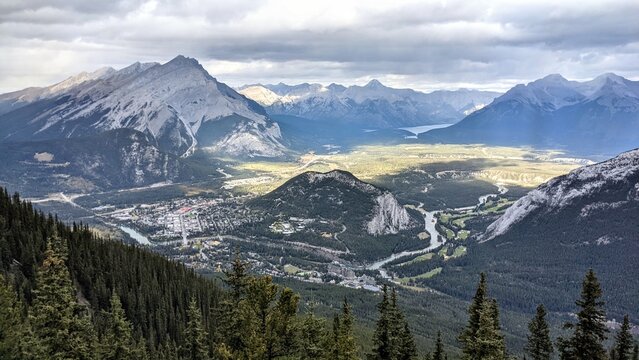 A view of the town of Banff from the top of Sulphur Mountain in Alberta. On a cloudy day, the sun beams through the clouds and lights up the terrain nearby, creating contrasts in shadows and colour.