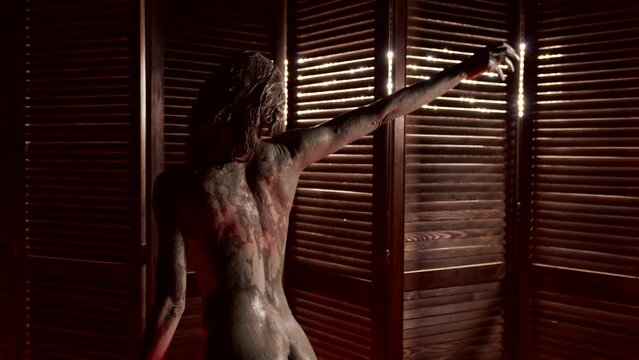 naked female body covered by mud in spa salon, sexy woman is dancing, rear view