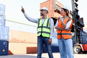 Portrait of two workers wearing safety vest and helmet discuss at logistic shipping cargo container yard, senior engineer man point and ask opinion of beautiful young woman colleague at workplace.