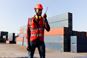 Portrait of African American young engineer worker man wearing safety bright neon red color vest and helmet, talking to a colleague with walkies-talkie at logistic shipping cargo container yard.