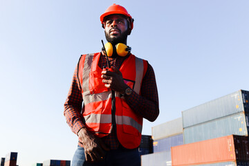 Portrait of African American young engineer worker man wearing safety bright neon red color vest and helmet, talking to a colleague with walkies-talkie at logistic shipping cargo container yard.