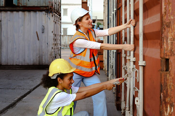 Two beautiful young worker engineer women wear safety vest and helmet, try to open shipping container door at logistic cargo yard. African American and Caucasian girls work together as same workplace.