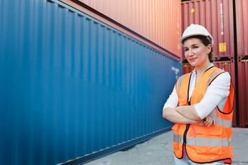 Portrait of happy smiling young beautiful woman engineer boss with blonde hair wearing safety vest and helmet, standing with confidence arm crossed at logistic shipping cargo container yard.