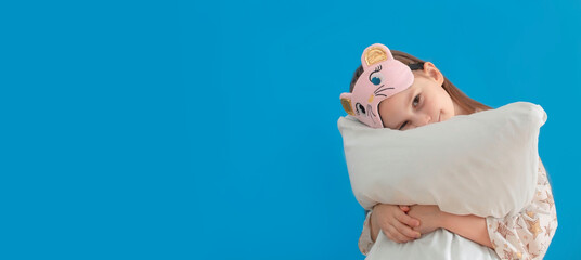Banner. Portrait of a positive cheerful child in an eye mask, hugging her big soft pillow on a blue...