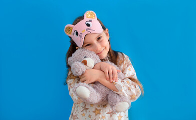 Portrait of a positive cheerful child in an eye mask, hugging a teddy bear on a blue background,...