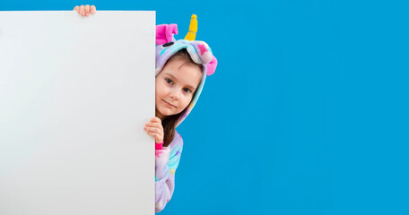 baby girl in a cozy blue unicorn costume stands behind a white empty banner or an empty advertising board on a blue one. A pretty girl is holding an empty banner