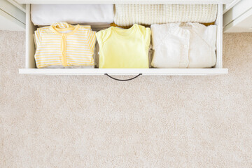 Folded baby clothes in white drawer box at nursery room at home. Closeup. Empty place for text on light carpet background. Top down view.