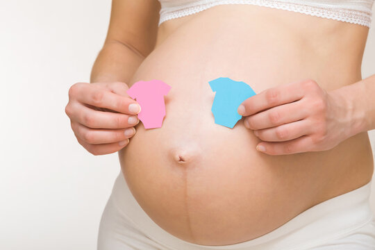 Hands holding shapes of pink and blue bodysuits on young adult pregnant woman big naked belly. Guessing future baby gender. Baby expectation. Closeup. Isolated on light gray background.