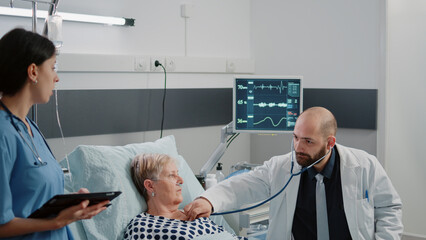 Doctor using stethoscope for heartbeat measurement on sick patient in hospital ward bed. Medic doing healthcare consultation with aged woman while nurse with tablet giving assistance