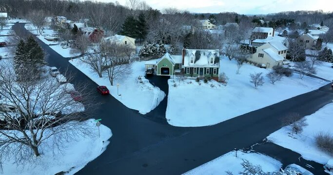 Green Cape Cod home covered in winter snow. Car passes by at night with headlights on. Aerial establishing shot.