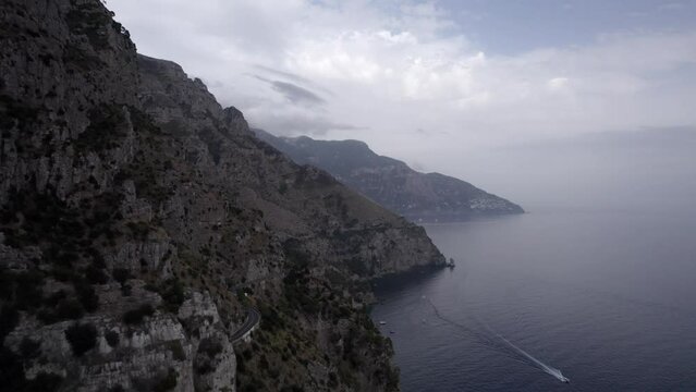 Drone video over the Amalfi Coast near Positano flying over the cliff with a frontal plane advancing in the direction of Positano.
Cloudy sky shows boats and vehicles and a boat sailing.