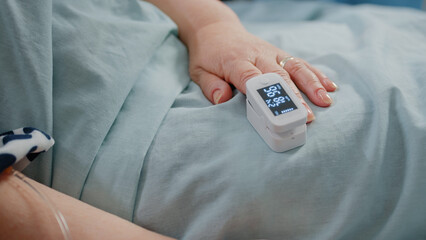 Close up of hand of aged patient with oximeter in bed. Senior person having medical tool on finger for oxygen saturation and pulse pressure measurement. Pensioner with sickness