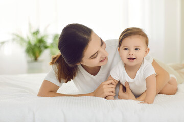 Fototapeta na wymiar Smiling young mom and small baby infant lie relax in bed at home playing together. Loving caring mother with little newborn kid in bedroom. Happy motherhood concept. Childcare and maternity.