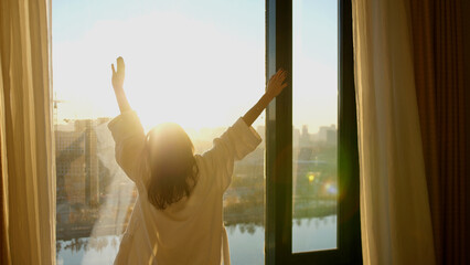 Young woman in white bathrobe open curtains in bedroom after waking up. Lady stretching with rising hands, looking out panoramic window enjoying sunrise in early morning © emaria