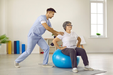Male doctor with help of fitball trains elderly female patient in rehabilitation clinic. Young male rehabilitator in uniform helps old woman perform exercises while sitting on blue fitball.