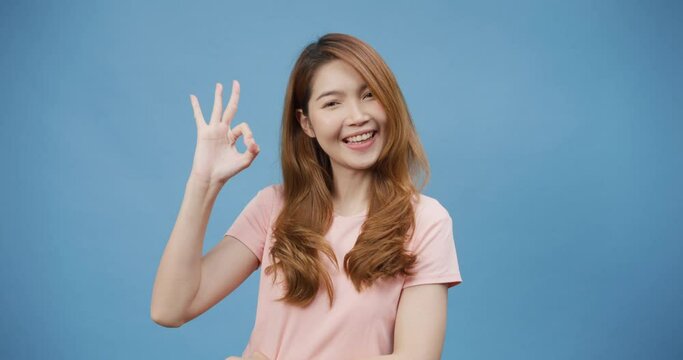 Attractive cheerful youth Asia lady wear casual pink cloth with okay sign good choice finger, look at camera smile stand isolate over blue background. Recommend product, copy space concept.
