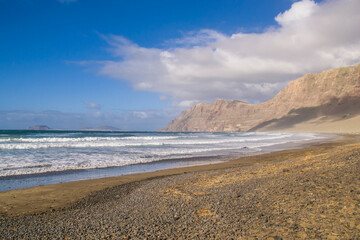 Fototapeta na wymiar A view of the beach (Playa) in Famara, Lanzarote. At the back of the beach are the high cliffs (Risco) and on the horizon the island of La Graciosa.