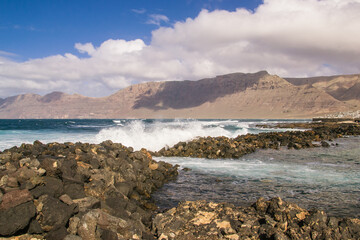 Fototapeta na wymiar Waves breaking against the rocks protecting the beach at Famara in Lanzarote. In the background are the cliffs, Ricso de Famara.