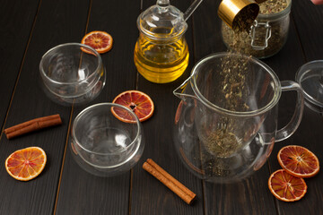 a glass teapot for brewing, two double-walled bowls, honey in a jar, a jar of natural tea, slices of dried red orange, cinnamon sticks are placed on a wooden table