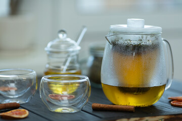 on a wooden table there is a glass teapot for brewing, two bowls with double walls, honey in a jar, a jar of natural tea, slices of dried red orange, cinnamon sticks on the table, the kettle is filled
