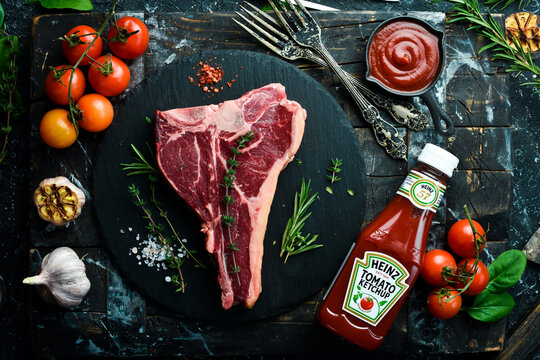 UKRAINE, LVIV - February 10, 2022: A bottle of Heinz Ketchup and Raw T-bone or porterhouse beef meat Steak on black wooden background. Top view. On a black stone plate.