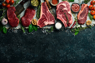 Variety of raw aged beef steaks: t-bone, tomahawk, striploin, tenderloin, new york steak for grilling with spices on stone background. On a black stone background.