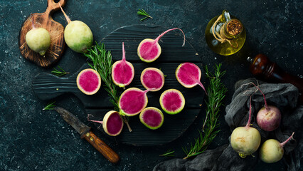 Pink watermelon radish lying on a couple of whole light green watermelon radishes. On a black stone background.