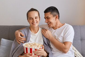 Portrait of smiling joyful wife and husband sitting on sofa with pop-corn and remote control, watching comedy or funny show, laughing happily and hugging, couple spending time together.