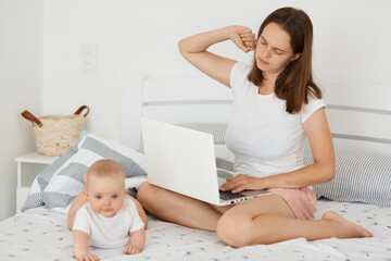 Portrait of tired exhausted woman sitting on bed with her infant daughter and working online, holding laptop and stretching hands, spending long hours working online.