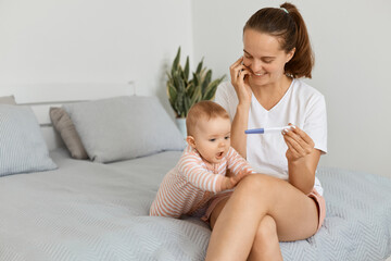 Fototapeta na wymiar Indoor shot of smiling happy woman wearing white t shirt and shorts sitting on bed with her baby daughter and holding pregnancy test with positive result, calling to husband to say good news.
