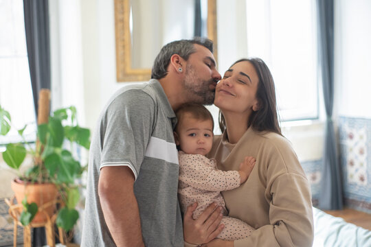Portrait of happy mid adult Caucasian family with little child standing together at home. Woman holding baby son and man kissing his wife. Family at home concept
