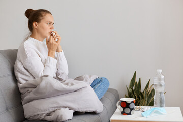 Profile portrait of sick woman being wrapped in blanket sitting on sofa with handkerchief and nasal...