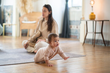 Cute baby boy crawling in room by his mother. Young woman sitting on floor in nursery with her...