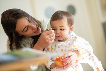 Obraz na płótnie Canvas Happy young mother feeding baby son with spoon and holding apple in morning. Little child having breakfast with mom at home. Motherhood and childcare concept