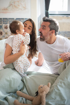 Happy Family Having Fun In Bed At Home. Woman Nursing Baby Boy, Man Making Face In Bedroom. Family At Home Concept