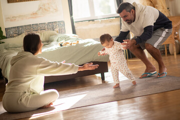 Happy parents teaching baby son to walk in bedroom. Mid adult couple with little child spending time together at home in morning. First steps, childcare, parenthood concept