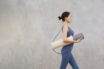Asian girl with a yoga mat and sportswear against a gray textured wall