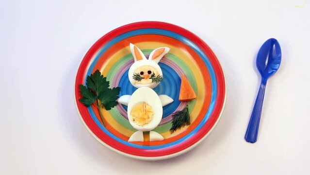 Easter breakfast idea for kids. Bunny rabbit made of boiled eggs on multicolored plate, white background, top view. Fun food for children.