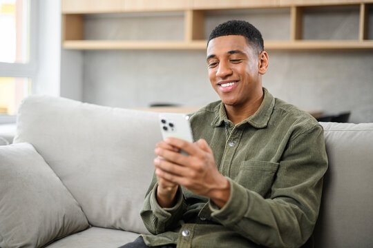 Focused Latin Man Sitting On The Sofa At Home And Holding Smartphone. Hispanic Guy Reading News Online, Spending Leisure Time In Social Networks. Male Texting And Chatting Online