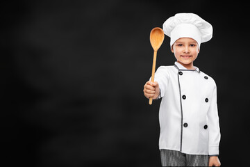 Naklejka premium cooking, culinary and profession concept - happy smiling little girl in chef's toque and jacket with wooden spoon over black chalkboard on grey background