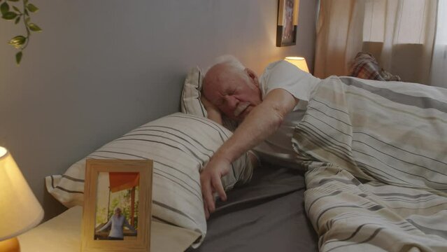 Aged Caucasian man with mustache waking up alone in his bed, moving hand on empty side of bed in search of deceased wife, looking at her picture in morning