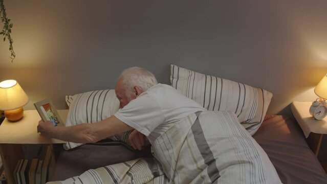 High-angle of senior Caucasian man with mustache waking up alone in his bed, looking at picture of wife in frame in morning
