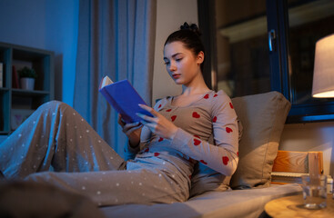 people, bedtime and rest concept - teenage girl reading book in bed at home at night