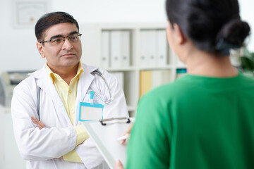 Pensive serious general practitioner with empty badge on labcoat talking to nurse filling medical form